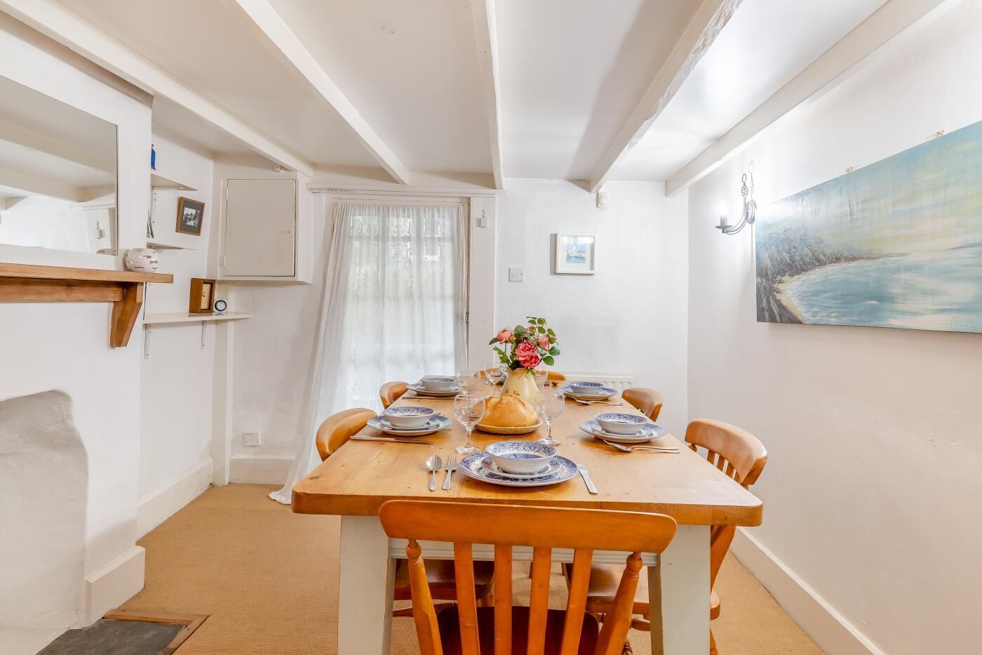 Uma sala de jantar com mesa de madeira posta para seis, paredes brancas, um quadro na parede e uma janela com cortinas leves.