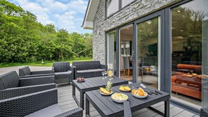 An outdoor patio with wicker furniture, a dining table set with food and drinks, and a view of the interior through large glass doors. - 4 Bed in Retorrick Mill (oc-t28899) (Saint Mawgan)
