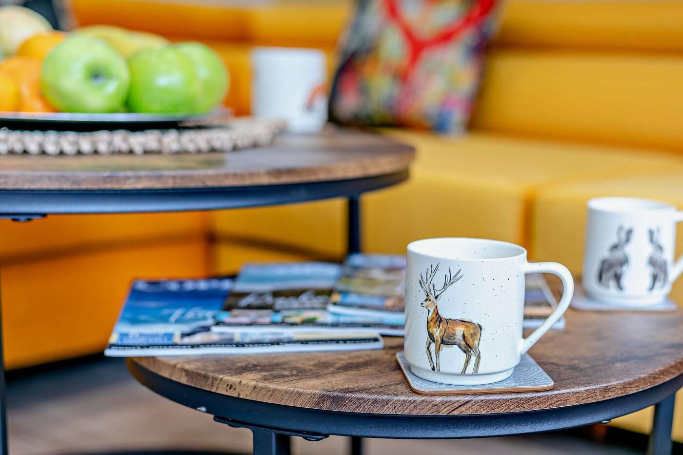 Une table en bois sur laquelle se trouvent une tasse ornée d’une illustration de cerf, une assiette de pommes et des magazines.