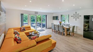 A modern living room with a large orange sofa, a dining area with a wooden table and chairs, and a kitchen with a refrigerator. - 4 Bed in Retorrick Mill (oc-w28897) (Saint Mawgan)