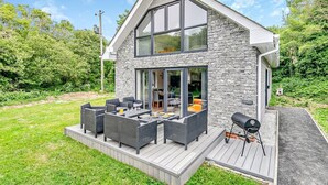 A modern house with a stone facade, large windows, and a deck with outdoor furniture. - 4 Bed in Retorrick Mill (oc-w28897) (Saint Mawgan)