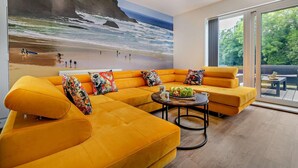 A modern living room with a large beach mural, a yellow sofa set, and a dining area with a view of the outdoors. - 4 Bed in Retorrick Mill (oc-w28897) (Saint Mawgan)