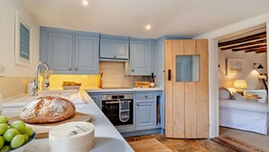 A modern kitchen with blue cabinets, a wooden door, and a dining area with a white sofa and lamp. - 3 Bed in Dunwich (oc-l18684) (Dunwich)