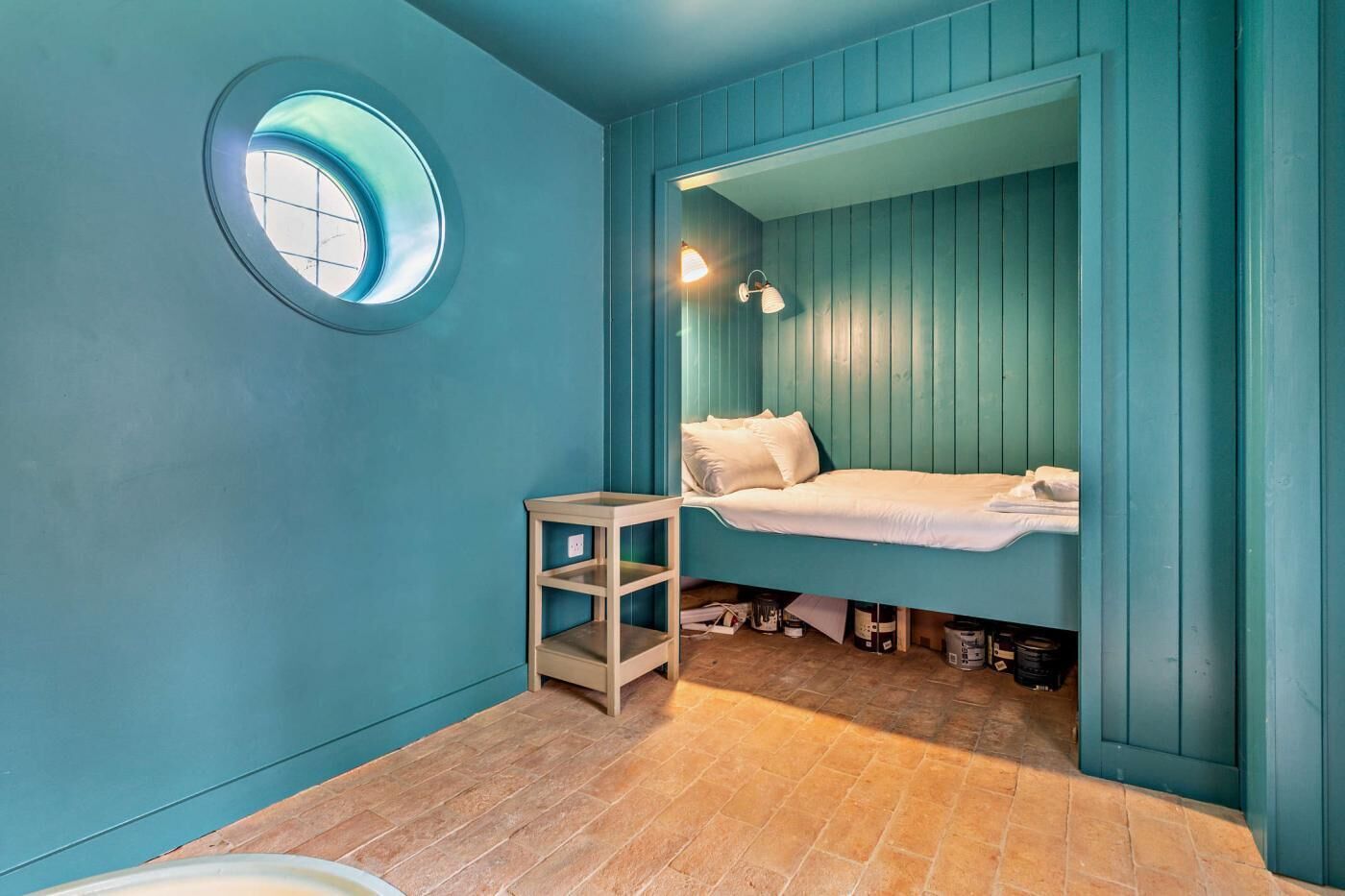 A small, cozy room with teal walls, a round window, a bed, and a small wooden table.
