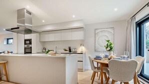A modern kitchen with white cabinetry, a central island, and a dining area with wicker chairs. - 2 Bed in Harlyn Bay (oc-s28921) (Harlyn Bay)