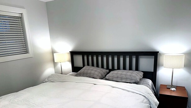 A neatly made bed with a white comforter and two pillows, flanked by two bedside tables with lamps, in a room with light gray walls. - Charming 2 BR 1BA Basement suit in White Rock with WiFi (White Rock)