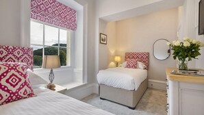 Een slaapkamer met twee bedden, een raam met gordijnen met patroon, een bedlampje, een nachtkastje, een kaptafel en een vaas met bloemen.