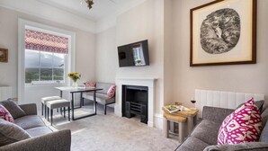 Een woonkamer met een open haard, een televisie, een eethoek met een tafel en stoelen, en een groot raam met decoratieve gordijnen.