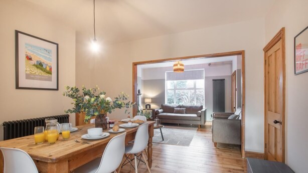 Un coin repas avec une table en bois, des chaises blanches et un vase de fleurs. On aperçoit un canapé et une lampe au fond.