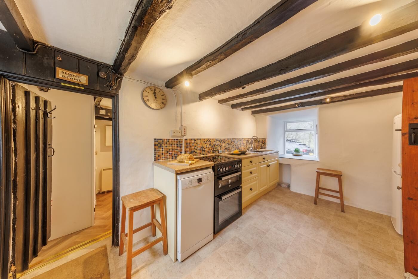 A kitchen with a tiled backsplash, wooden beams, a clock, and a window.