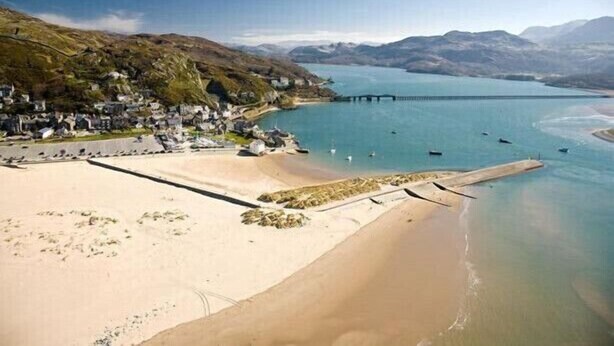 Een kustlandschap met een zandstrand, een rustige baai en een pier die in het water reikt.