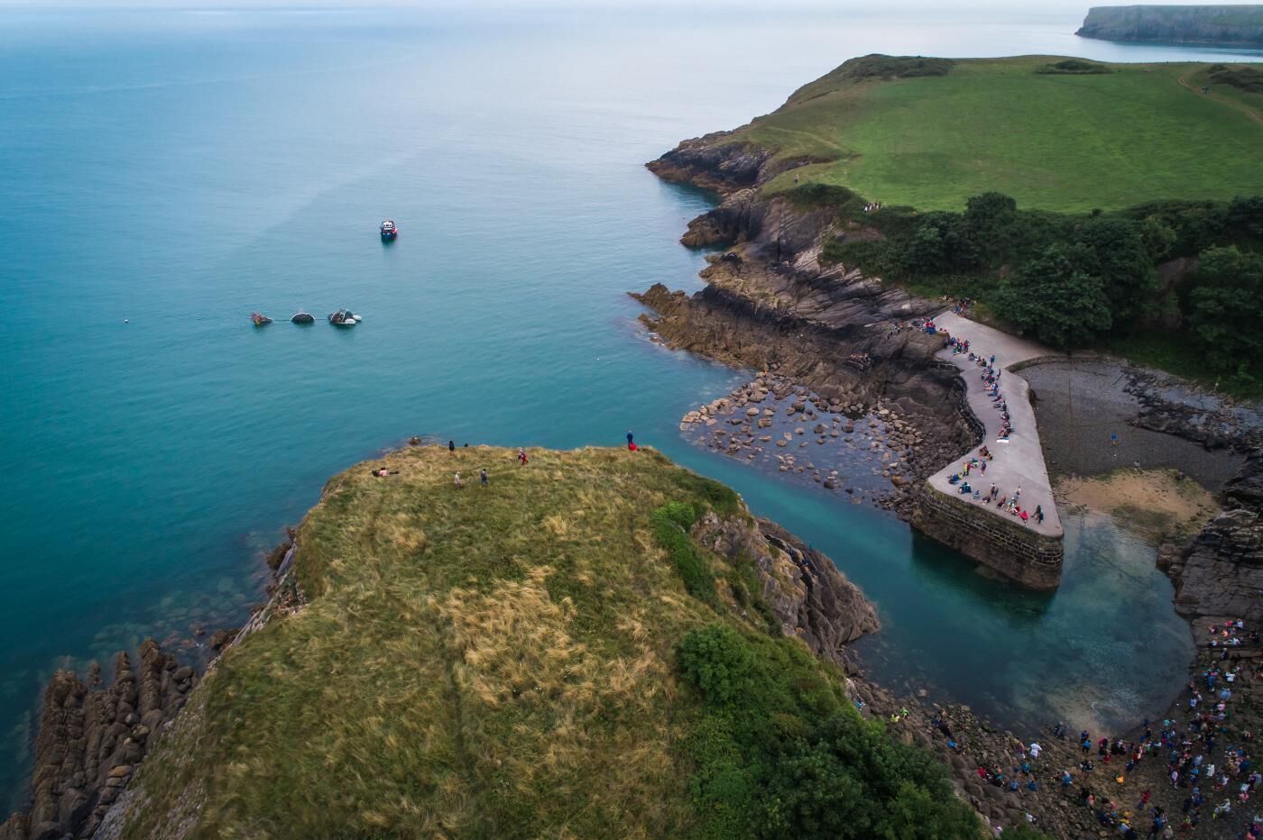 An aerial view of a coastal area with a stone pier extending into the sea, surrounded by rocky cliffs and grassy terrain.