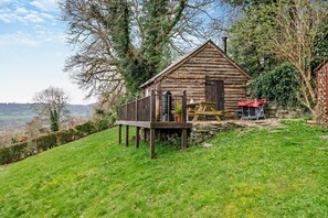 A rustic log cabin with a deck, surrounded by greenery and a stone wall.