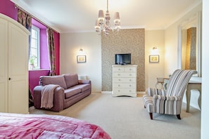 A bedroom with a bed, a sofa, a dresser, a chair, a television, and a chandelier.