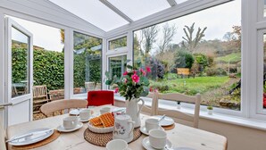 A conservatory with a table set for breakfast, a red chair, and a view of a garden. - 3 Bed in Axmouth (oc-t28930) (Axmouth)