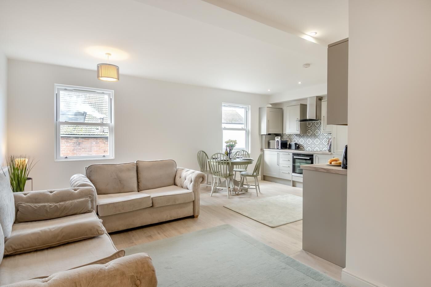 A modern living room with a beige sofa, a dining table with chairs, and a kitchen area with cabinets and appliances.