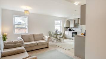 A modern living room with a beige sofa, a dining table with chairs, and a kitchen area with cabinets and appliances.