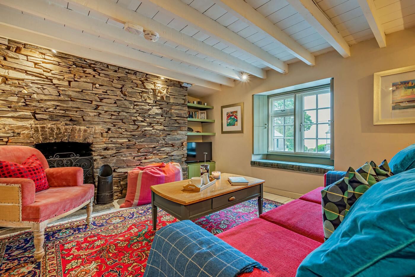 A cozy living room with a stone wall, a fireplace, a large window, and colorful furnishings.
