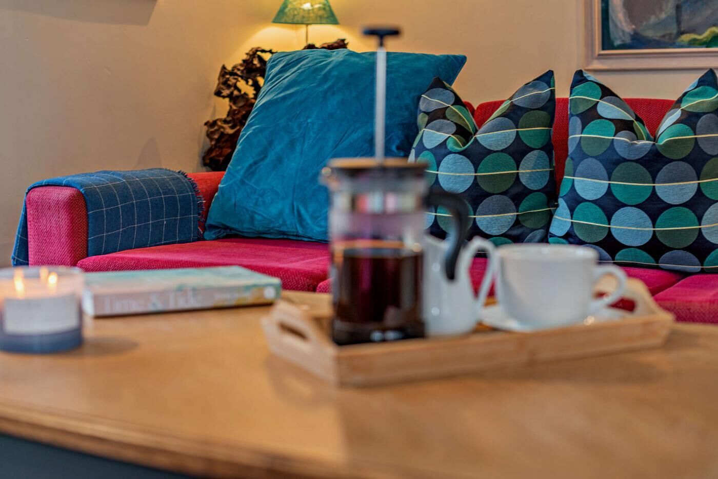 Un espacio de sala acogedor con una preparación para café, incluyendo una cafetera y tazas en una bandeja, un libro y una vela sobre una mesa de madera.