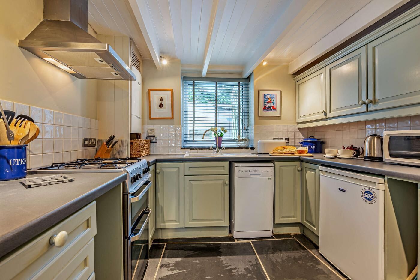 A compact kitchen with a gas stove, a dishwasher, and a microwave. There are cabinets and a window with blinds.