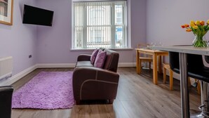 A living room with a sofa, a dining table, a TV mounted on the wall, and a window with blinds. - 1 Bed in Tenby (oc-w28984) (Tenby)