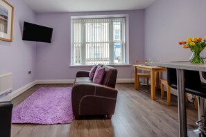 A living room with a sofa, a dining table, a TV mounted on the wall, and a window with blinds. - 1 Bed in Tenby (oc-w28984) (Tenby)