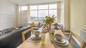A dining area with a long table set for breakfast, a sofa, and a view of the outdoors through large windows. - 2 Bed in Tenby (oc-b28989) (Tenby)
