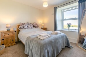 A neatly made bed with a grey blanket, two bedside tables with lamps, a window with curtains, and a view of the outdoors. - 1 Bed in Four Lanes (oc-f28972) (Four Lanes)