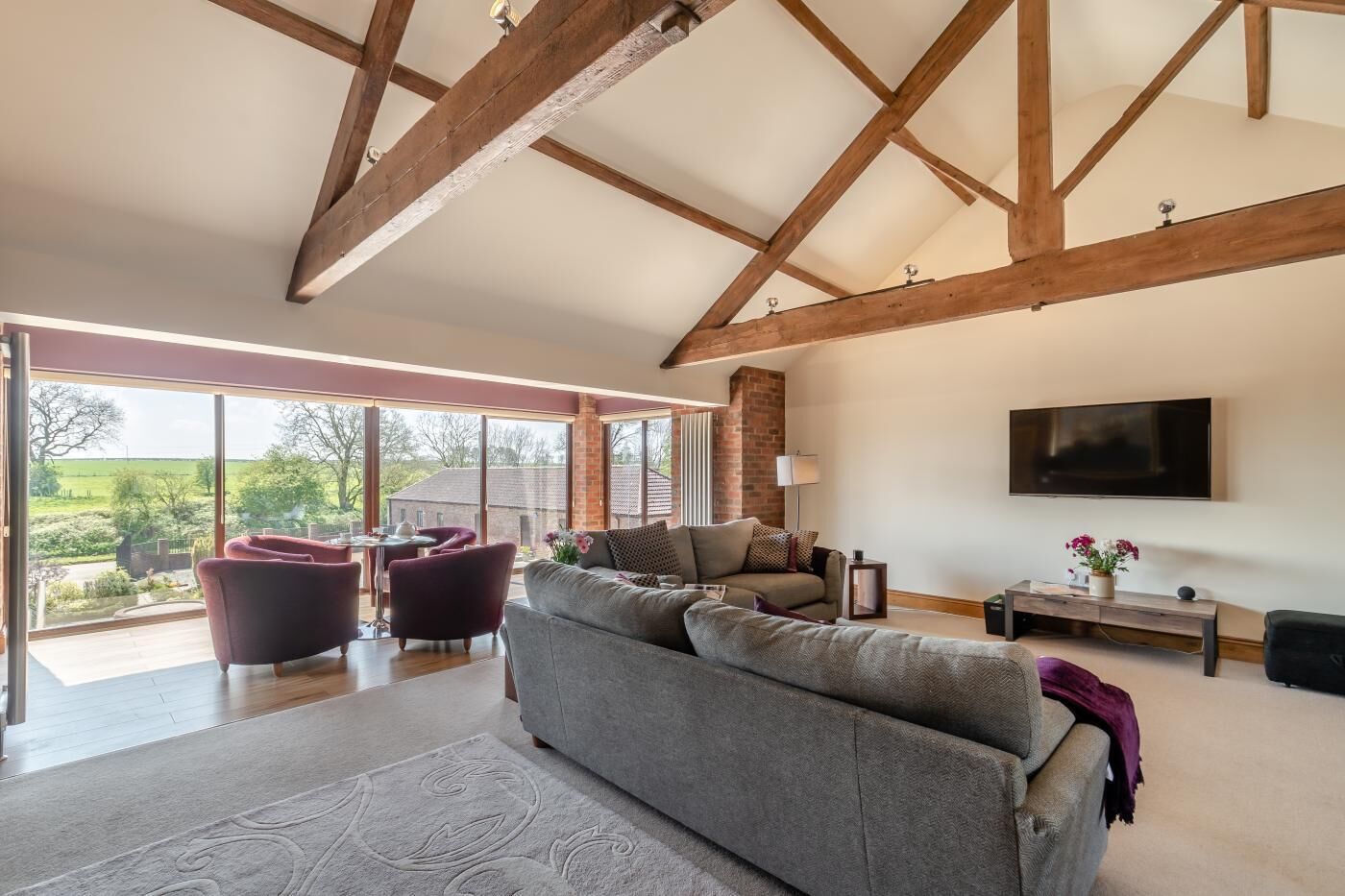 Ein geräumiges Wohnzimmer mit hohen Decken, großen Fenstern, einem grauen Sofa und einem Flachbildfernseher.