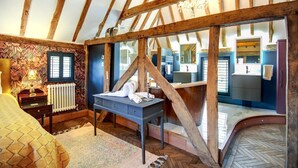Un dormitorio con vigas de madera a la vista, una cama, un escritorio y un baño visible a través de un ventanal.