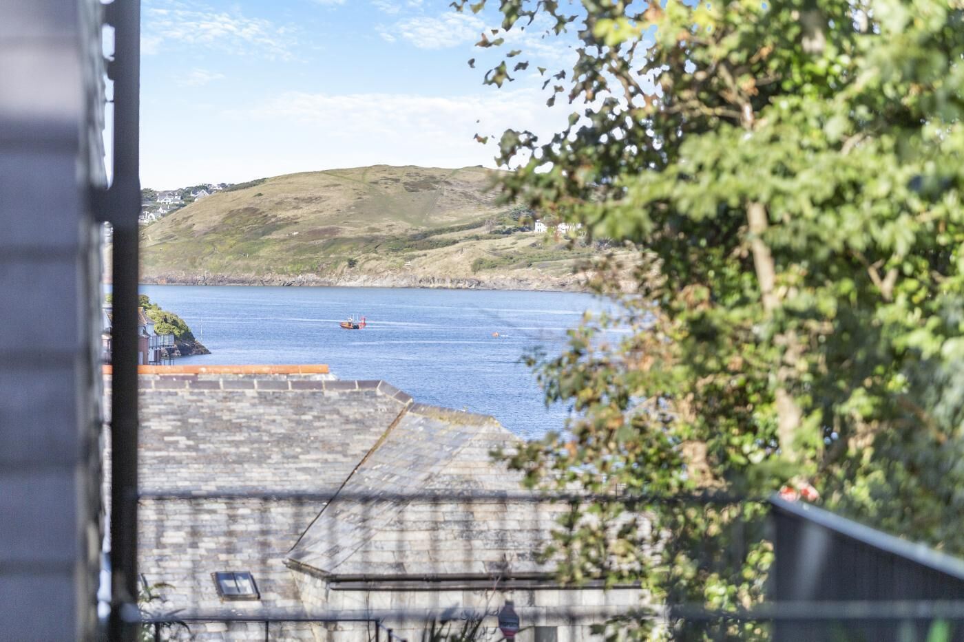 Blick auf die Küste mit einem Boot auf dem Wasser, einer Steinmauer und Grünpflanzen im Vordergrund.