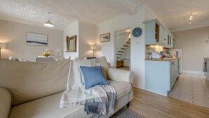 Ein Wohnzimmer mit einem beigen Sofa, einem blauen Kissen und einer weißen Tagesdecke. Es gibt eine Küchenzeile mit hellblauen Schränken und einer Uhr an der Wand.