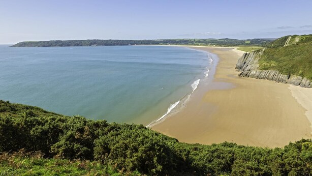 Eine Küstenlandschaft mit sandigem Strand, tiefblauem Meer und einer grasbewachsenen Klippe.