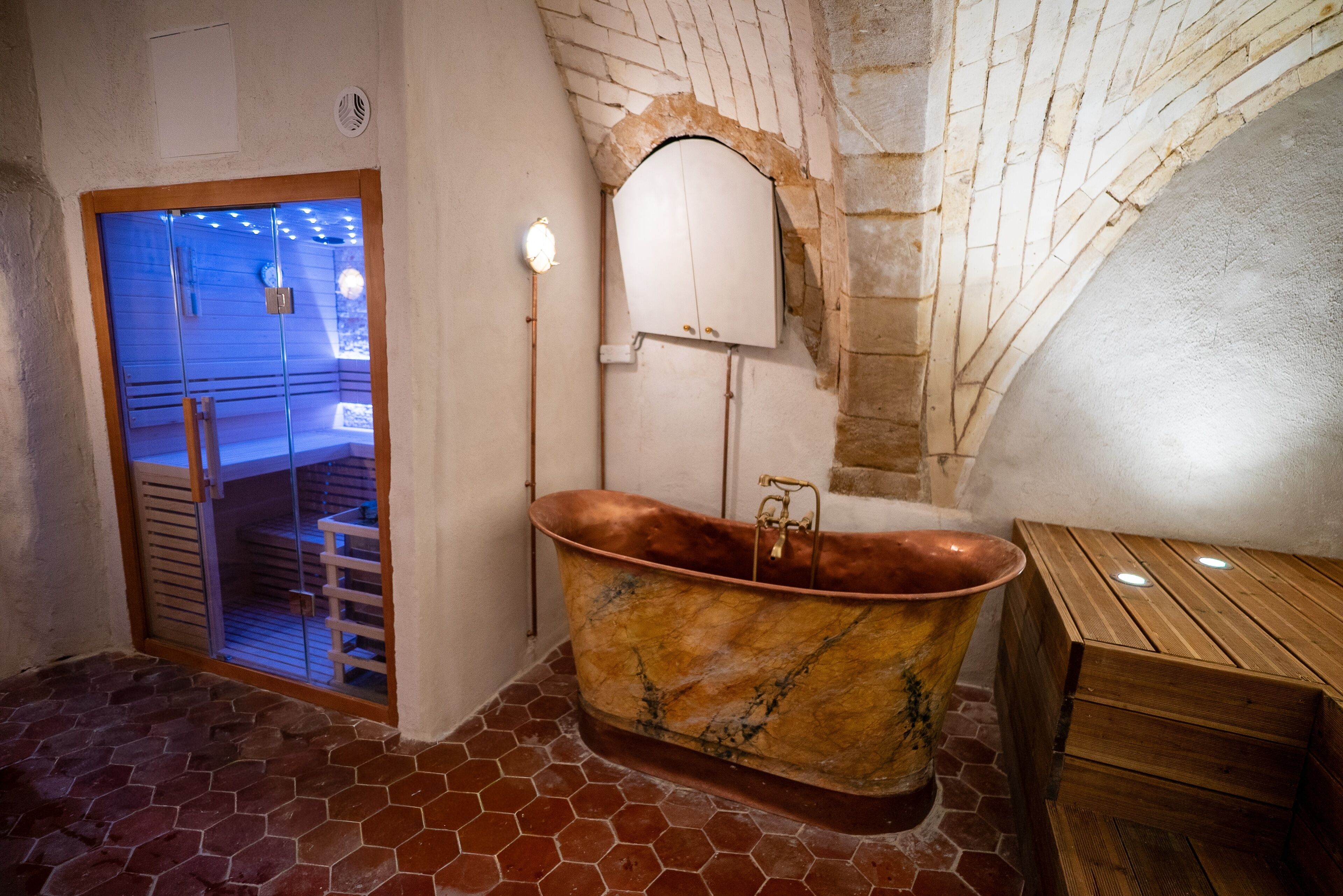 A room with a large copper bathtub, a wooden bench, a stone wall, and a glass door leading to a seating area.