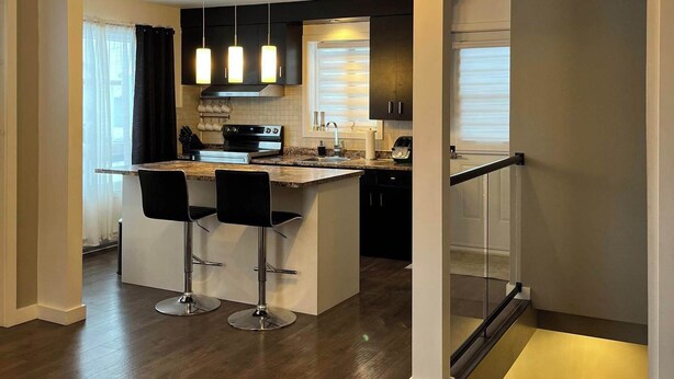 Una cocina moderna con una isla central, taburetes de bar, iluminación colgante y vistas al área de estar.
