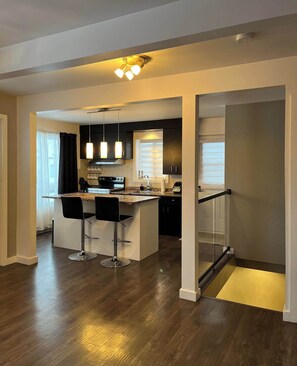 A modern kitchen with a central island, bar stools, pendant lighting, and a view of the living area. - Cottage de style loft · 2 chambres · Deschambault-Grondines (Grondines)