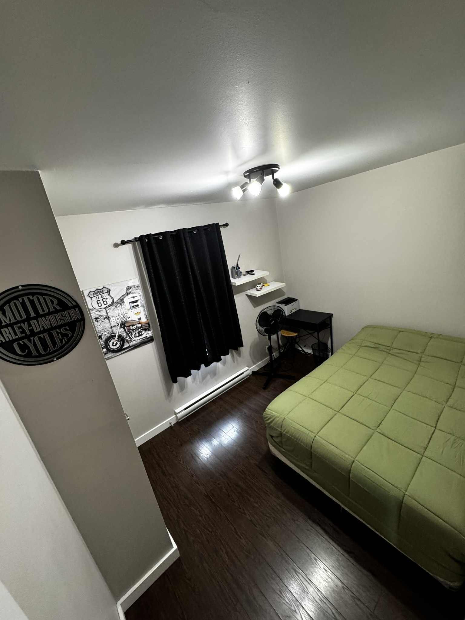 A small, well-lit bedroom with a bed, a desk, and a chair. There is a window with a black curtain, a wall-mounted light fixture, and a wall decoration.