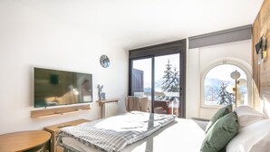 Quarto com cama, televisão, escrivaninha e janela com vista para montanhas.