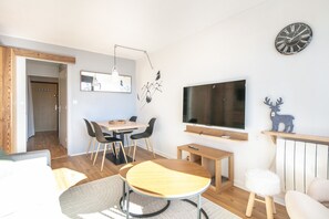 A modern living room with a dining area, a TV mounted on the wall, and a wooden-framed window. - Le Roche Mantel - Appart 4p à Alpe d'Huez (Huez)