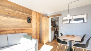 Uma sala de estar moderna com uma parede de destaque em madeira, um sofá cinzento, uma área de jantar com mesa de madeira e cadeiras pretas, e uma cozinha ao fundo.
