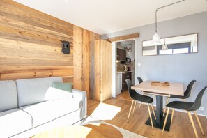 A modern living room with a wooden accent wall, a grey sofa, a dining area with a wooden table and black chairs, and a kitchen in the background. - Le Roche Mantel - Appart 4p à Alpe d'Huez (Huez)