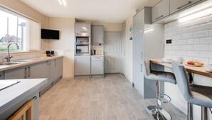 A modern kitchen with grey cabinets, a built-in oven, a refrigerator, and a dining area with a table and chairs. - 3 Bed in Bacton (oc-l2294) (Bacton)