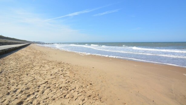 A wide sandy beach with gentle waves, a clear sky, and a concrete barrier on the left. - 3 Bed in Bacton (oc-l2294) (Bacton)
