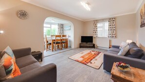 A living room with a sofa, a coffee table with fruit, a TV, and a dining area with chairs and a table. - 3 Bed in Bacton (oc-l2294) (Bacton)