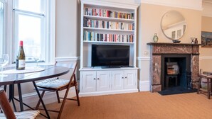 Een woonkamer met een boekenkast, een open haard, een televisie en een eethoek met een tafel en stoelen.