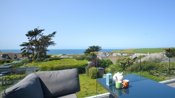 Un balcon avec un canapé gris, une table en verre et une vue sur la mer et les maisons.