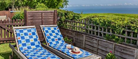 Two wooden lounge chairs with blue patterned cushions, a wooden table with a hat and a bottle, and a wooden fence with greenery.