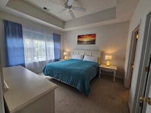 Ein Schlafzimmer mit einem Bett, zwei Nachttischen, einem Deckenventilator, einem Fenster mit Vorhängen und einem Bild an der Wand.
