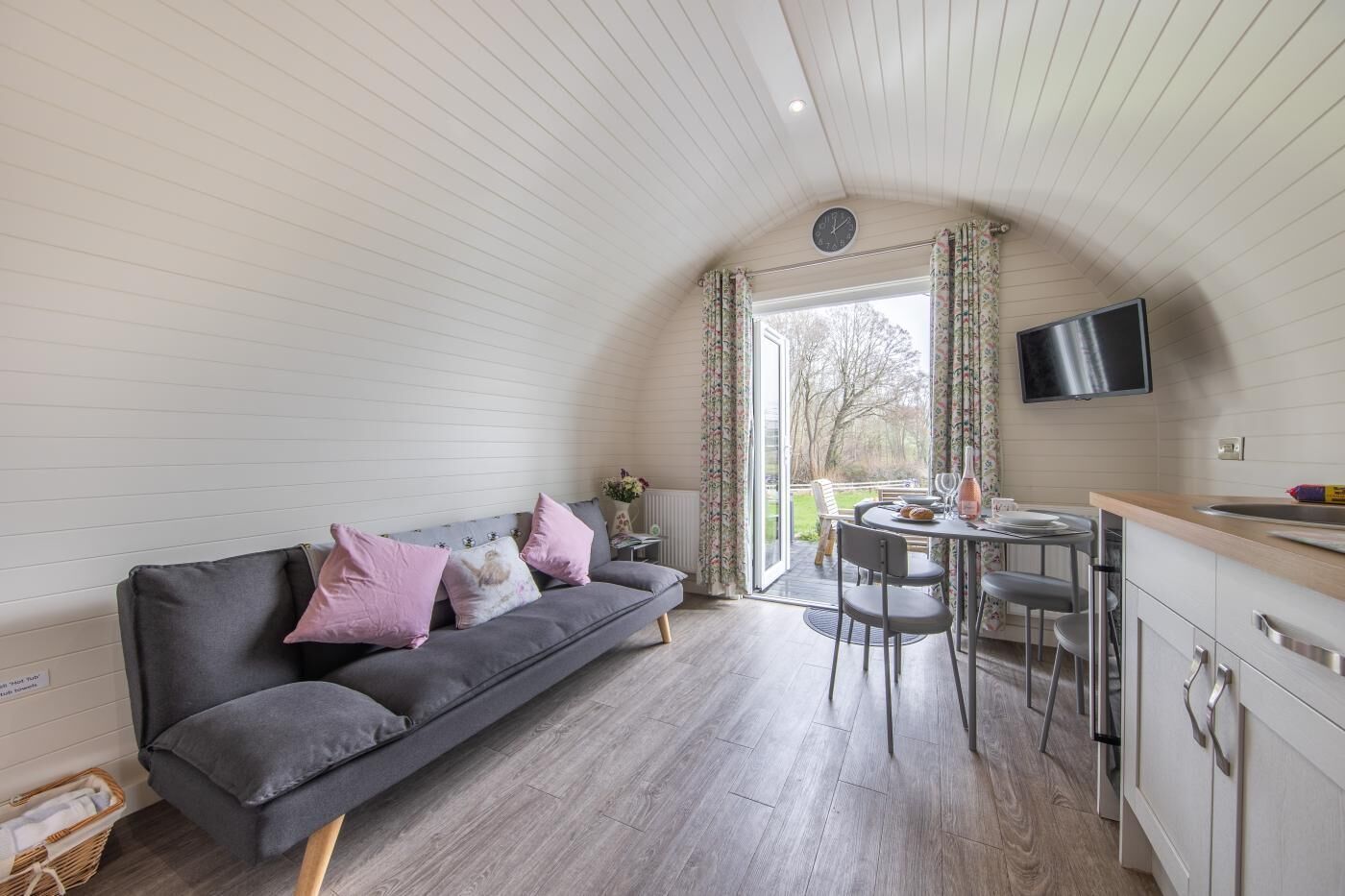 A cozy room with a grey sofa, a dining table with chairs, a TV mounted on the wall, and a window with curtains.