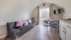 Ein gemütliches Zimmer mit grauem Sofa, einem Esstisch mit Stühlen, einem an der Wand befestigten Fernseher und einem Fenster mit Vorhängen.